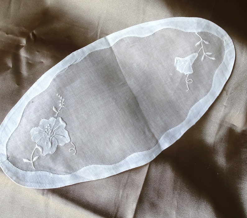May include: White oval-shaped linen table runner with embroidered floral designs. The runner has a scalloped edge.