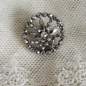 May include: A silver-toned button with a decorative design of a flower with a circular frame. The button is made of small, faceted beads.