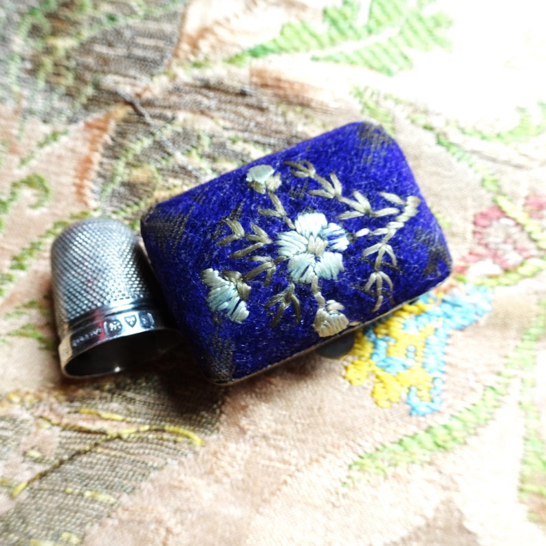 May include: A silver thimble with an inscription and a small blue velvet box with a floral embroidery design.