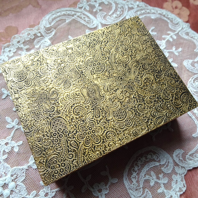 May include: A gold box with a black and gold paisley pattern. The box is made of a textured material and has a slightly raised design.