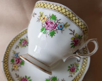 Puede incluir: Juego de taza y platillo blancos con un diseño floral con flores rosas, amarillas y azules. El borde y los bordes están adornados con un patrón geométrico amarillo y dorado. La taza tiene un asa curvada.