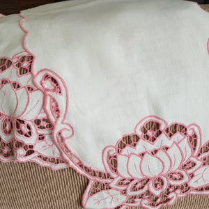 May include: White linen table runner with pink embroidered floral design. The design features delicate flowers and leaves.