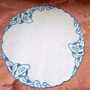 May include: A white, round doily with a scalloped edge. The doily features a blue floral embroidery design around the edge, with stylized flowers and leaves. The embroidery adds a decorative touch to the classic white fabric.