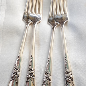 May include: Four silver forks with decorative floral patterns on the handles. The forks are displayed on a white surface, highlighting their polished finish and intricate details. A classic set for dining.