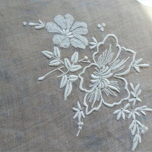 May include: White embroidered floral design on a sheer white fabric. The design features a large flower with smaller flowers and leaves.