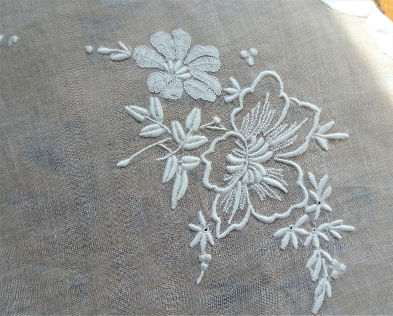 May include: White embroidered floral design on a sheer white fabric. The design features a large flower with smaller flowers and leaves.