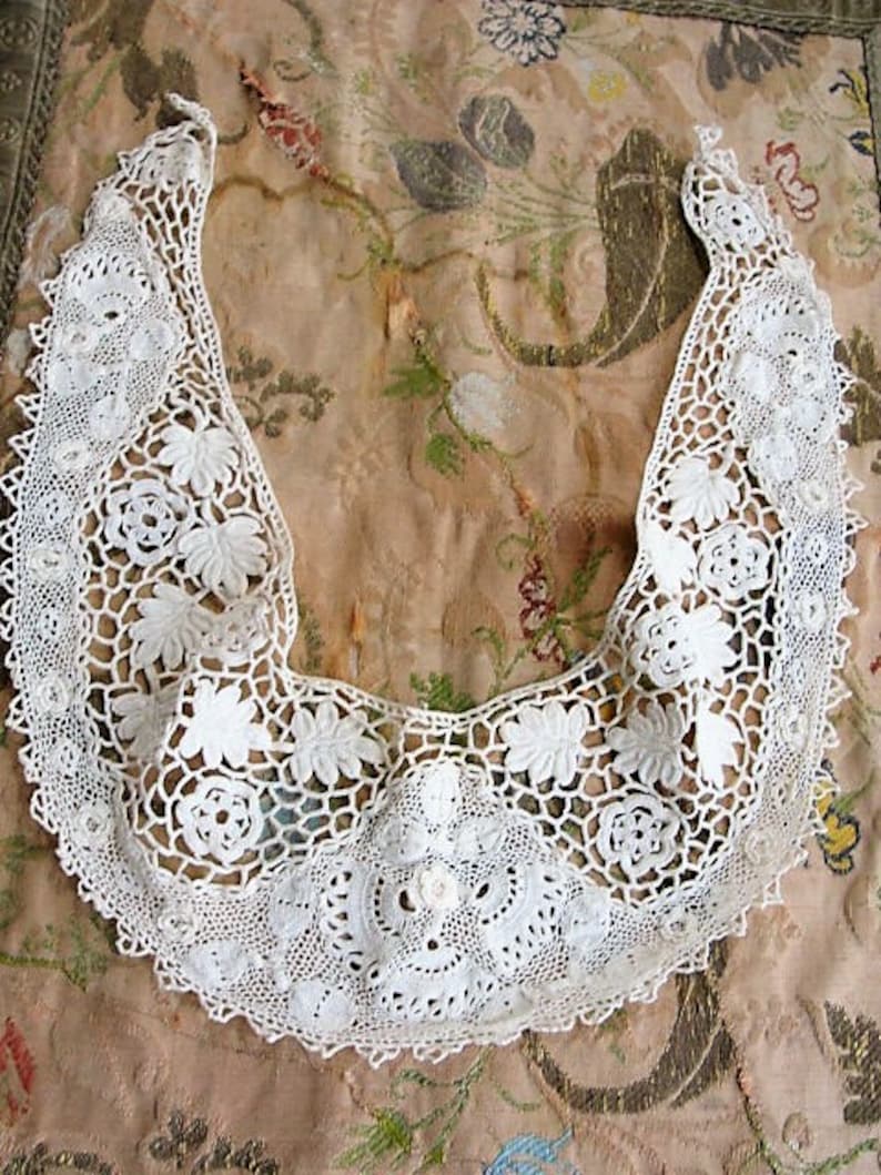 May include: A white crocheted lace collar with a scalloped edge and floral designs. The collar is laid out on a patterned fabric background.
