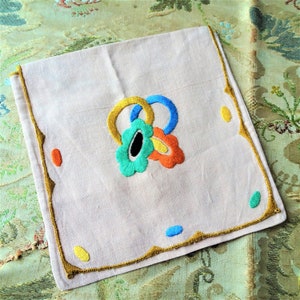 Puede incluir: Una servilleta de lino blanco con un borde bordado en oro y un diseño floral colorido en el centro. El diseño presenta colores azul, verde, amarillo y naranja.