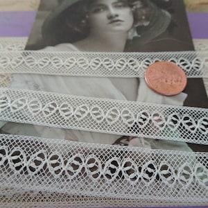 May include: White cotton lace trim with a delicate floral pattern. The lace is approximately 1 inch wide and is shown on top of a vintage postcard.