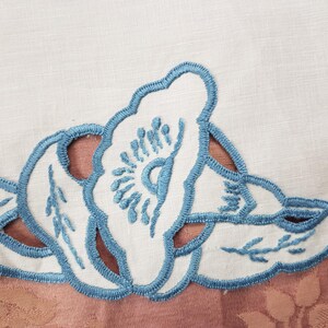 May include: Close-up of a white fabric with a floral design. The design features white cut-out flowers and leaves with light blue embroidered outlines. The background is a muted pink fabric with a floral pattern.