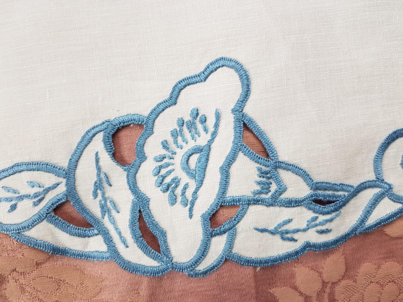 May include: Close-up of a white fabric with a floral design. The design features white cut-out flowers and leaves with light blue embroidered outlines. The background is a muted pink fabric with a floral pattern.