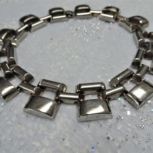 Puede incluir: Un collar de cadena de plata con eslabones grandes y rectangulares. El collar está hecho de metal pulido y tiene un diseño moderno y geométrico.