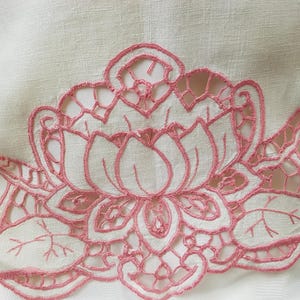 May include: Pink embroidered floral design on a white linen fabric. The design features a large flower with intricate details and leaves.