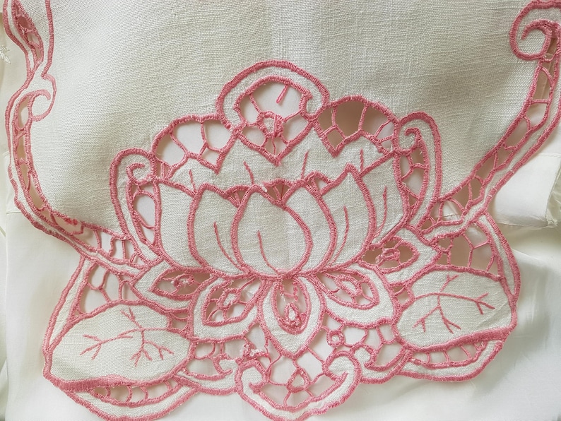 May include: Pink embroidered floral design on a white linen fabric. The design features a large flower with intricate details and leaves.