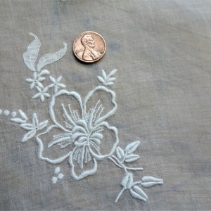 May include: White embroidered floral design on a sheer white fabric. The design features a large flower with smaller flowers and leaves surrounding it.