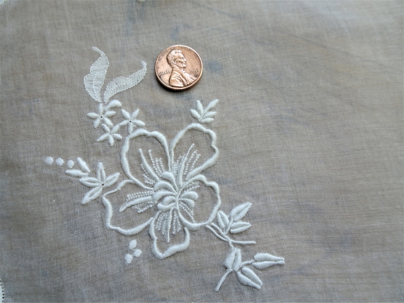 May include: White embroidered floral design on a sheer white fabric. The design features a large flower with smaller flowers and leaves surrounding it.