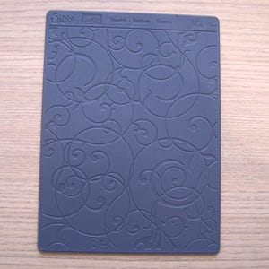 May include: A blue Sizzix embossing folder with a swirling flourish design. The folder has the text "Flourish" and other text. The design is a repeating pattern of curved lines and circles, creating a textured effect.
