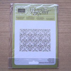 May include: A clear plastic package containing a Big Shot die-cutting machine plate. The plate features a detailed, brown, floral, and scrollwork design. The package includes text in multiple languages, including "Big Shot" and "Shown at 50%."