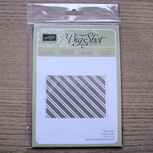 May include: A package containing a Big Shot die-cutting embossing folder. The folder features a diagonal striped pattern in white and gray. The packaging includes text that reads "Shown at 50%" and "Stampin' Up!"