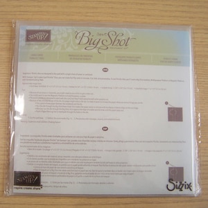 May include: A clear plastic package containing a white sheet of paper with text and graphics. The text includes "Stampin' Up!" and "Big Shot". The package is on a light brown wooden surface.