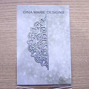 May include: A silver metal die cut in the shape of a half-circle with a detailed lace design. The die cut is in a clear plastic package with the text "GINA MARIE DESIGNS" printed on the front. The background is a light gray with a subtle pattern.