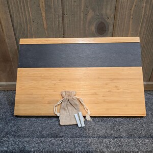 May include: A bamboo serving board with a dark gray slate insert. A small burlap bag with a drawstring closure and two white chalk sticks are in front of the board. The board is on a gray surface.