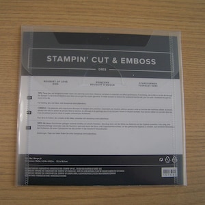 May include: A clear plastic package containing Stampin' Cut & Emboss dies. The package has the text "Stampin' Cut & Emboss" and "Bouquet of Love" printed on it. The package is on a wooden surface.