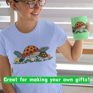 K&ouml;nnte beinhalten: Ein hellblaues T-Shirt mit einem Motiv von drei Pilzen in Braun-, Orange- und Gelbt&ouml;nen. Die Pilze sind von gr&uuml;nen Bl&auml;ttern umgeben. Der Text "Great for making your own gifts!" ist unten im Bild in Gr&uuml;n gedruckt.