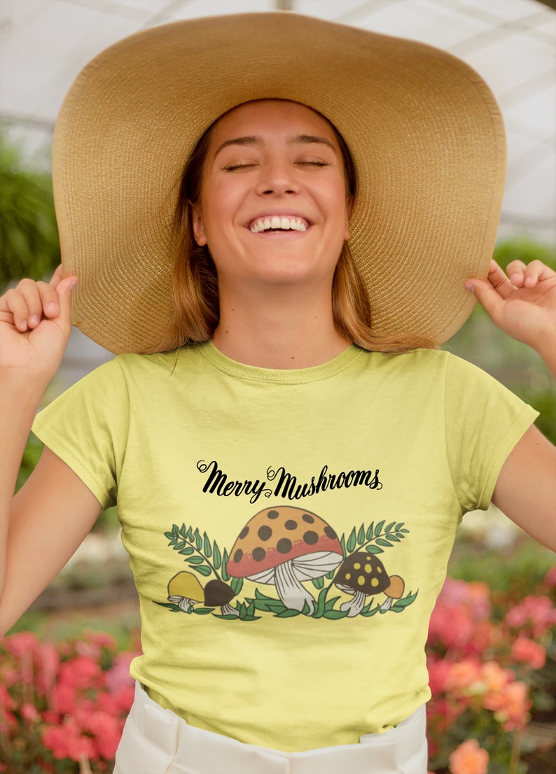 K&ouml;nnte beinhalten: Ein gelbes T-Shirt mit einem Aufdruck von Pilzen und dem Text "Merry Mushrooms".