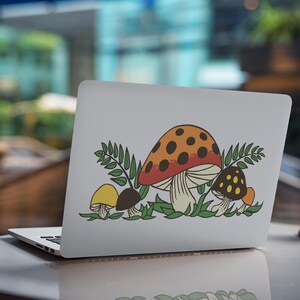 K&ouml;nnte beinhalten: Ein wei&szlig;er Laptop mit einem Aufkleber von drei Pilzen in einer Waldlandschaft. Die Pilze sind rot mit wei&szlig;en Punkten, gelb und braun mit wei&szlig;en Punkten. Der Aufkleber ist von gr&uuml;nen Bl&auml;ttern umgeben.