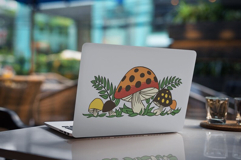 K&ouml;nnte beinhalten: Ein wei&szlig;er Laptop mit einem Aufkleber von drei Pilzen in einer Waldlandschaft. Die Pilze sind rot mit wei&szlig;en Punkten, gelb und braun mit wei&szlig;en Punkten. Der Aufkleber ist von gr&uuml;nen Bl&auml;ttern umgeben.