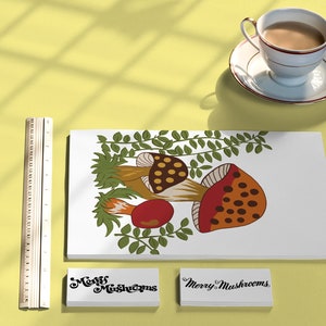 Puede incluir: Una tarjeta blanca con una ilustraci&oacute;n colorida de setas y hojas verdes. La tarjeta est&aacute; rodeada por una regla, una taza de t&eacute; y dos tarjetas blancas con el texto "Merry Mushrooms" en cursiva negra.