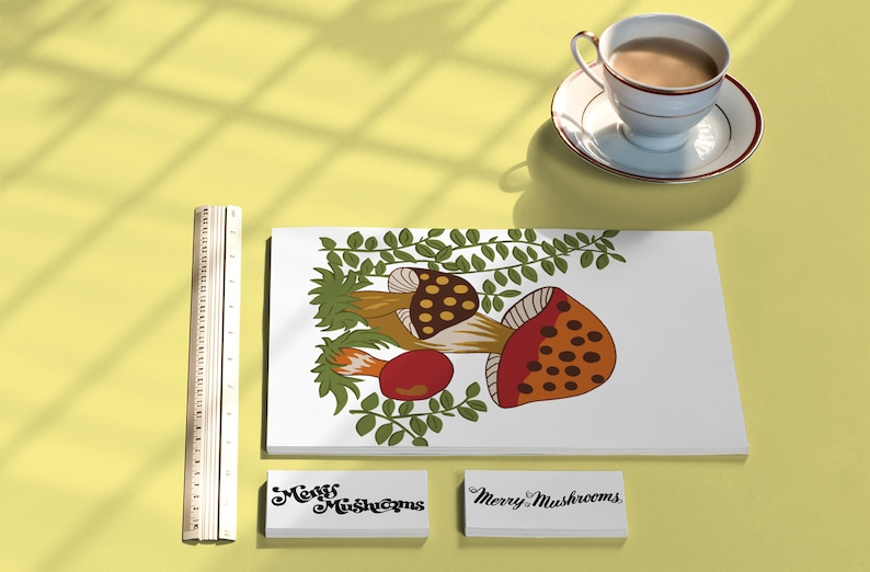 Puede incluir: Una tarjeta blanca con una ilustraci&oacute;n colorida de setas y hojas verdes. La tarjeta est&aacute; rodeada por una regla, una taza de t&eacute; y dos tarjetas blancas con el texto "Merry Mushrooms" en cursiva negra.