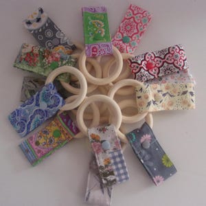 May include: A set of eight wooden teething rings with fabric loops attached. The loops are made of different patterned fabrics in shades of blue, green, pink, and yellow. Each loop has a button sewn on it.