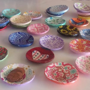 May include: A collection of small, round, shallow ceramic dishes featuring various colourful patterns, including floral, paisley, heart, and animal designs. Some dishes contain small items like coins or rings, while others are empty and arranged.