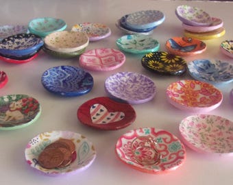 May include: A collection of small, round, shallow ceramic dishes featuring various colorful patterns, including floral, paisley, heart, and animal designs. Some dishes contain small items like coins or rings, while others are empty and arranged.