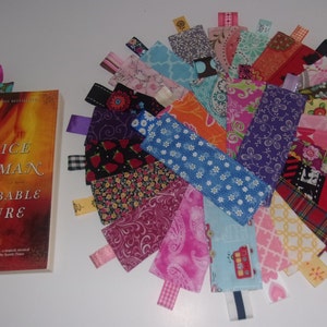 May include: A collection of 25 colorful fabric bookmarks with different patterns and designs. The bookmarks are arranged in a fan shape on a white surface. Each bookmark has a ribbon attached to the top.