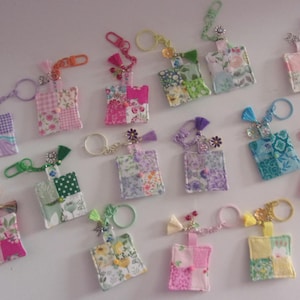 May include: A collection of colorful fabric keychains with floral and patchwork patterns. Each keychain features a metal ring and a decorative tassel or charm. The keychains are in various colors, including pink, green, and purple.