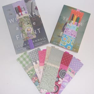 May include: A collection of fabric elastic pen holder bookmarks featuring various patterns including floral, paisley, polka dot, and gingham prints. Each bookmark has a lace trim detail and is designed to wrap around a book cover to hold pens.