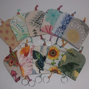 May include: A collection of fabric keychains with various patterns, including floral, geometric, and butterfly designs. Each keychain has a metal ring and a decorative tassel. The keychains are in shades of beige, blue, green, orange, and yellow.