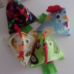 May include: Four colorful fabric chick-shaped toys. Each toy is a triangular pillow with a different pattern: crayons, bees, farm animals, and tractors. The toys have googly eyes and red felt combs.