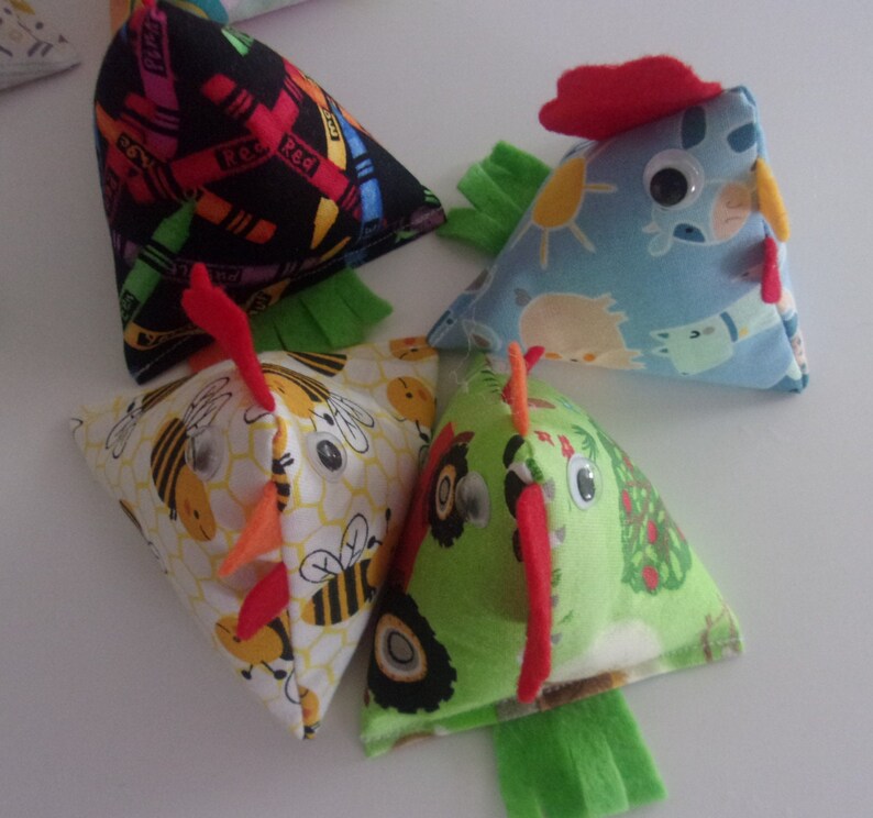 May include: Four colorful fabric chick-shaped toys. Each toy is a triangular pillow with a different pattern: crayons, bees, farm animals, and tractors. The toys have googly eyes and red felt combs.