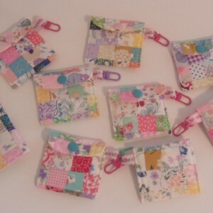 May include: A collection of nine small, square patchwork pouches with various floral and geometric patterns in pastel colors. Each pouch has a snap closure and a pink or purple clip.