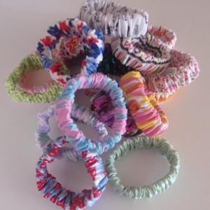May include: A collection of fabric hair scrunchies in various colors and patterns, including floral, striped, and solid designs. The scrunchies are gathered in a pile, showcasing different textures and prints such as blue, red, pink, green, and white.