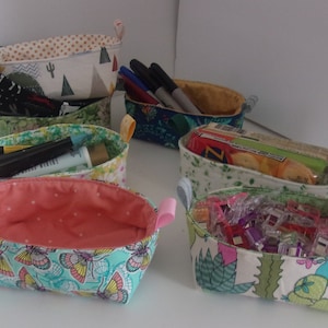 May include: Five fabric storage baskets with different patterns and colors. The baskets are filled with various items, including pens, pencils, snacks, and hair clips.