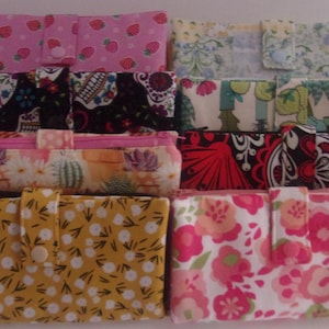 May include: A collection of eight rectangular fabric pouches arranged in two columns. Each pouch features a unique pattern, including strawberries, floral prints, cacti, and geometric designs. Every pouch has a central snap closure tab.
