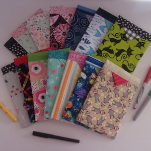 May include: A collection of colorful fabric pen cases, each with a unique pattern. Designs include floral, geometric, and novelty prints. Pens and markers are scattered around the cases, suggesting their use for writing or drawing.