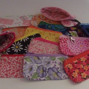 May include: A collection of small, colorful fabric zipper pouches in various floral and patterned designs. Some pouches are open, revealing coins or other contents. The pouches feature different colors, including pink, red, orange, and purple.