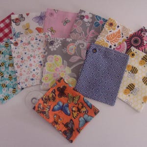 May include: A collection of fabric items with various patterns, including butterflies, bees, and floral designs. The fabrics feature colors like red, pink, blue, and yellow. Each item has a small hole at the top.