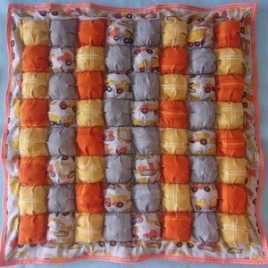 May include: A gray, yellow, and orange quilted baby blanket with a construction vehicle theme. The blanket is made of soft, plush fabric and has a quilted design with small squares. Each square features a different construction vehicle, such as a dump truck, bulldozer, and excavator. The blanket is perfect for a baby's nursery or playroom.